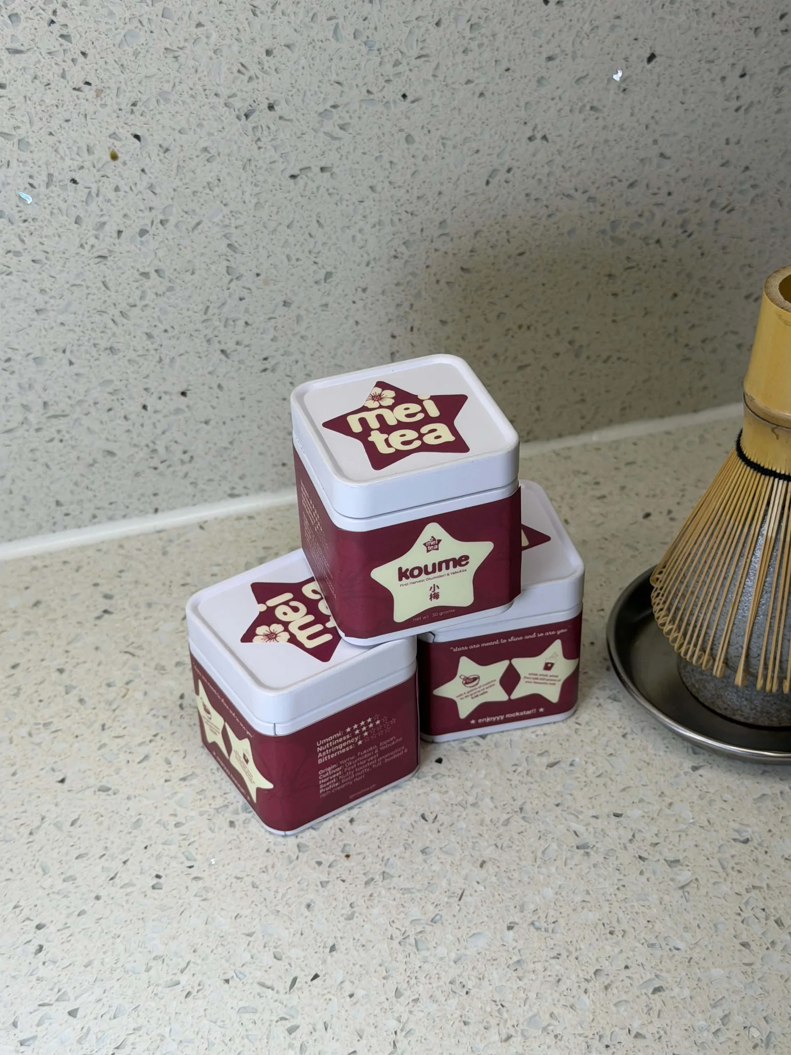 Koume tins with Mei Tea’s burgundy star labels, beside a bamboo whisk